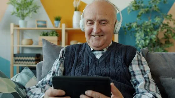 An elderly man sat on a sofa, he is reading something on a tablet whilst wearing big over ear headphones.