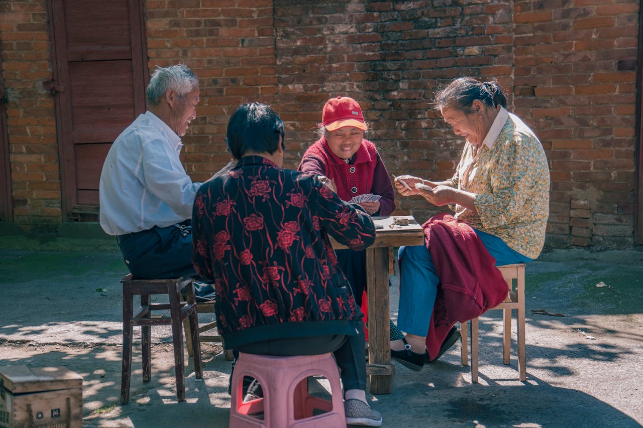 Group of people playing cards together outdoors beside a brick wall.