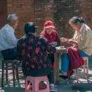 Group of people playing cards together outdoors beside a brick wall.