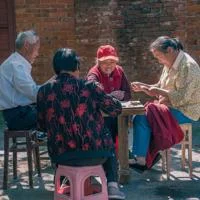 Group of people playing cards together outdoors beside a brick wall.