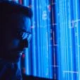 Close-up side profile of a person wearing glasses, looking at a blue digital interface with data lines and charts