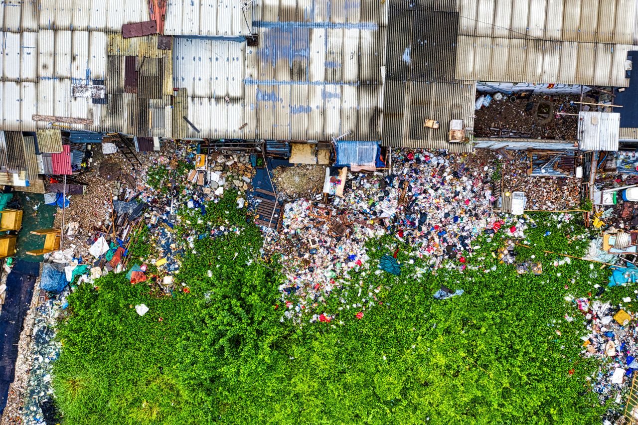 Aerial view of a large pile of mixed rubbish and waste materials beside a vibrant green area, with urban structures