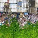 Aerial view of a large pile of mixed rubbish and waste materials beside a vibrant green area, with urban structures