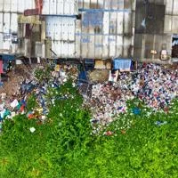 Aerial view of a large pile of mixed rubbish and waste materials beside a vibrant green area, with urban structures