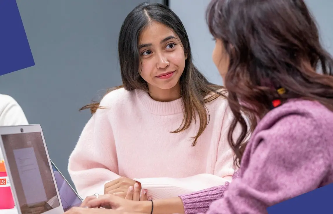  Two female students looking at each other. Their laptops open on the desk they're sharing.