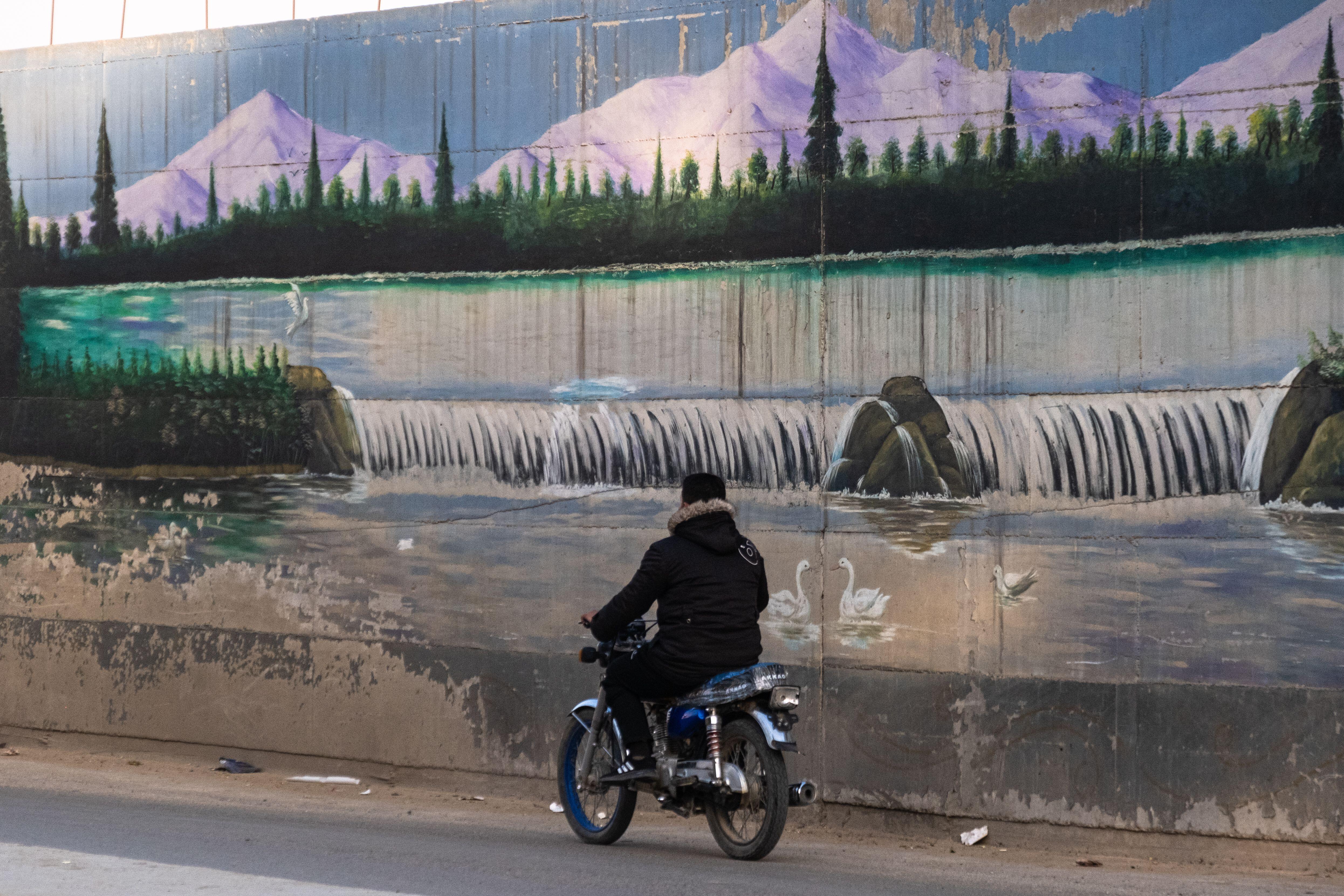 Man riding a motorbike alongside a utopian mural of mountains and waterfalls in Qamishli 
