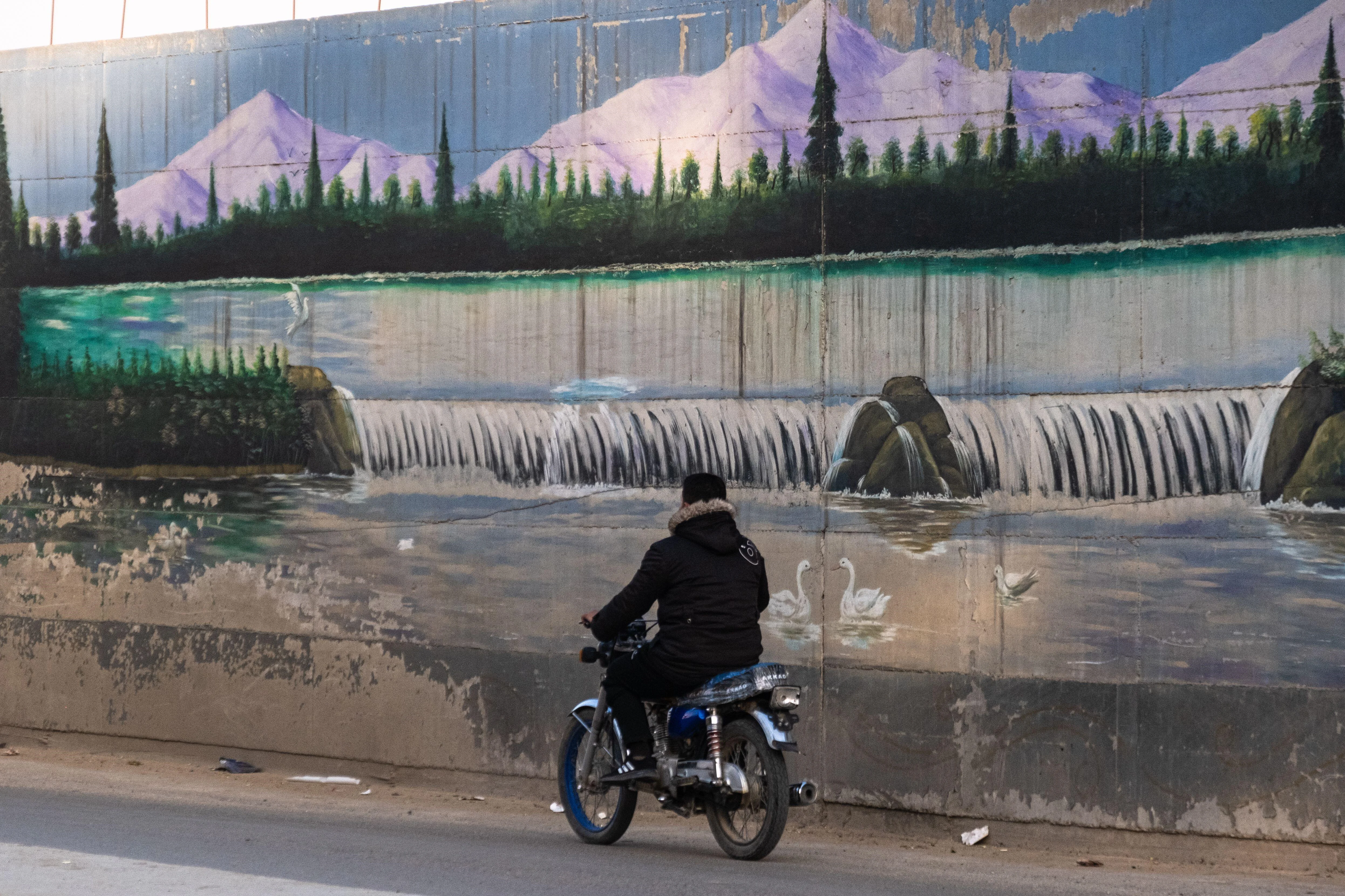 Man riding a motorbike alongside a utopian mural of mountains and waterfalls in Qamishli
