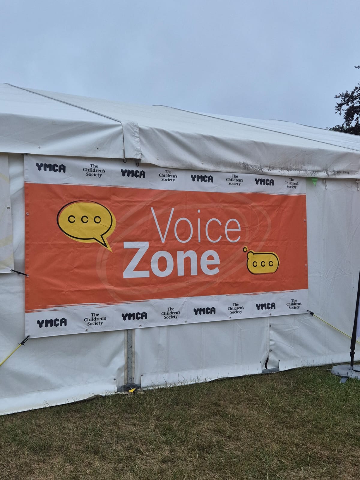 Orange banner on the side of a white outdoor tent reading “Voice Zone,” with speech bubble graphics and repeated YMCA and The Children’s Society logos.