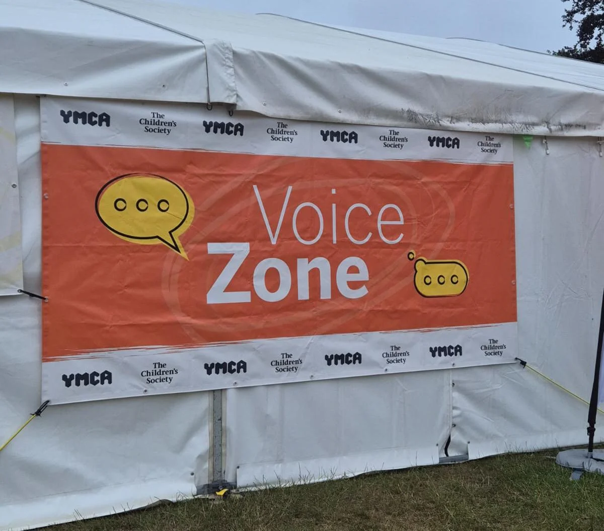 Orange banner on the side of a white outdoor tent reading “Voice Zone,” with speech bubble graphics and repeated YMCA and The Children’s Society logos.