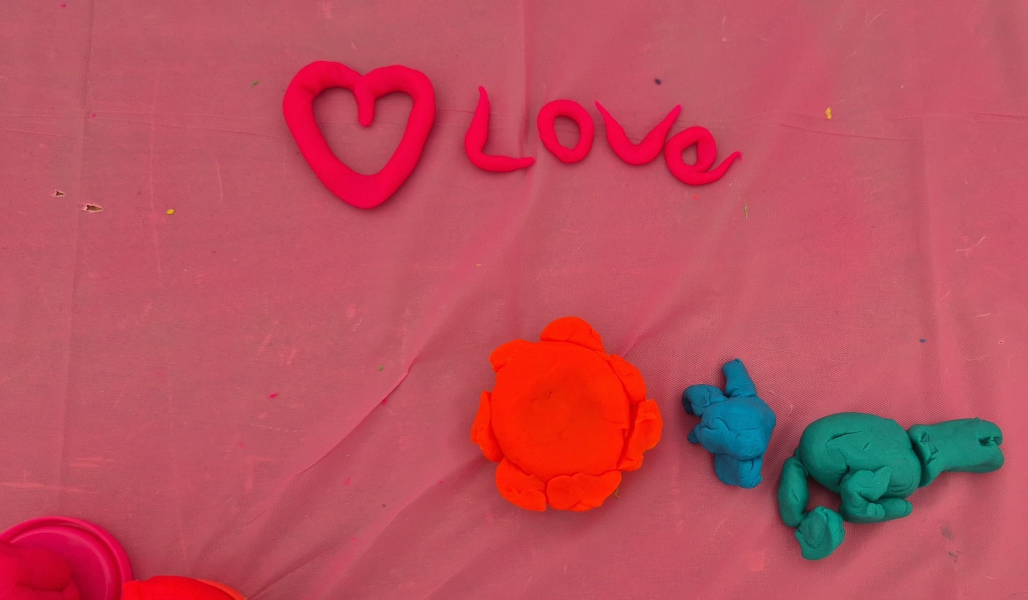 The word “Love” and a heart shape made from pink modelling clay on a pink tablecloth, with small colourful clay shapes below.