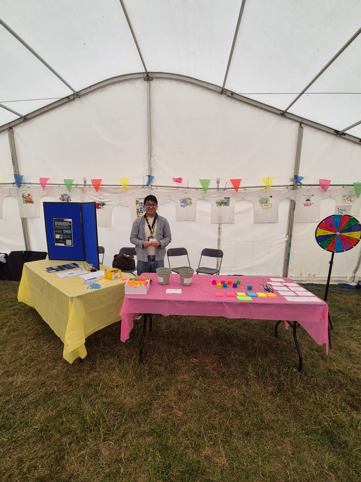A person standing behind two tables inside a white marquee tent. The tables are covered with yellow and pink cloths and display craft materials, coloured tokens, buckets, pens, sticky notes, and information leaflets. Colourful bunting hangs across the tent.