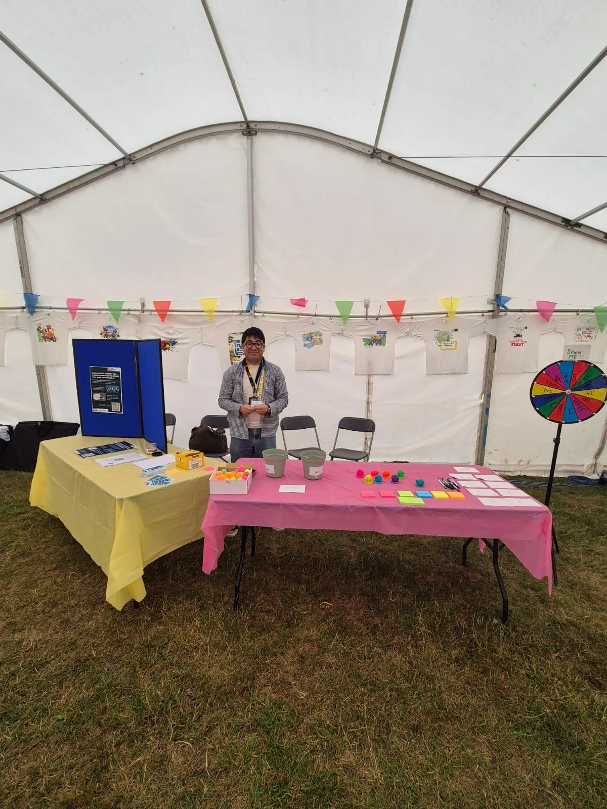 A person standing behind two tables inside a white marquee tent. The tables are covered with yellow and pink cloths and display craft materials, coloured tokens, buckets, pens, sticky notes, and information leaflets. Colourful bunting hangs across the tent.