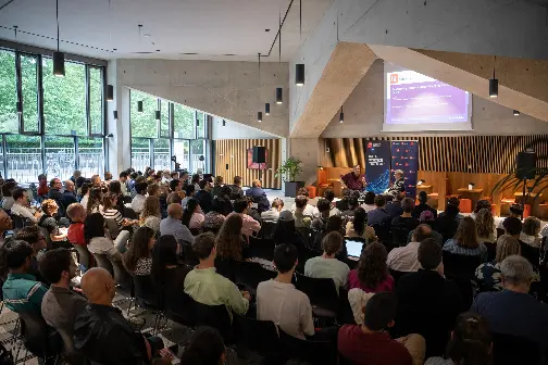 Audience attentively listens to a speaker in a modern conference room with large windows, concrete walls, and a banner.
