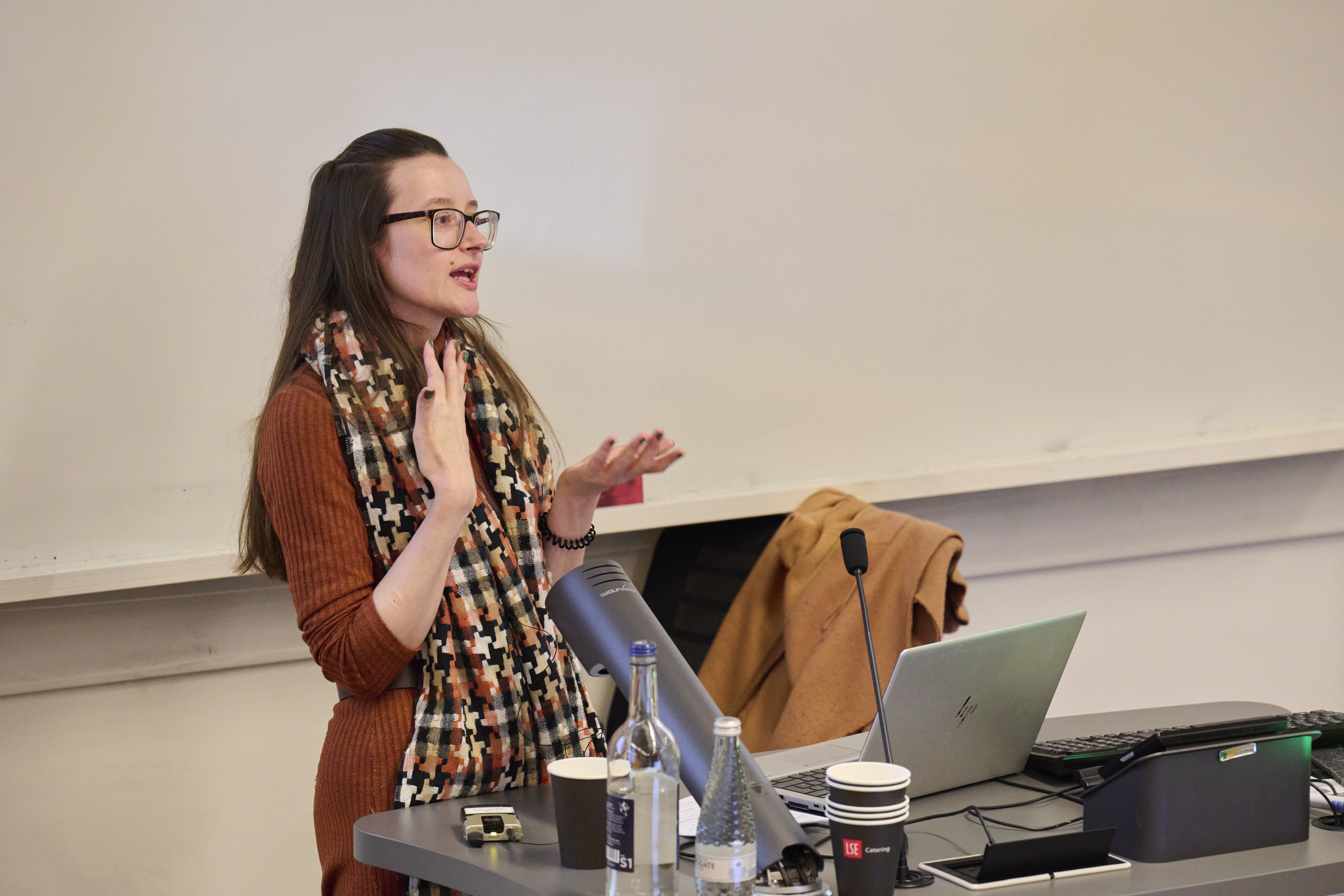 A woman gives a presentation. She wears glasses, has long dark hair and has a laptop open on the desk in front of her.