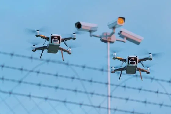 Two small drones in front of barbed wire. Three CCTV cameras are out of focus in the background.