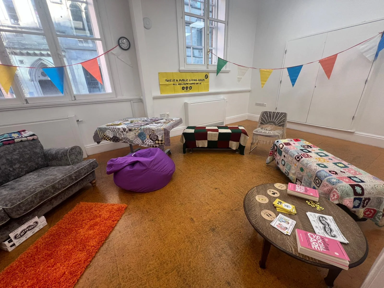 A bright community room set up like a shared living room, with mismatched sofas, chairs, a beanbag, and small tables covered with patterned cloths. Colourful triangular bunting hangs across the room. A yellow sign on the wall reads, “This is a public living room. All welcome, come on in.” Books and leaflets sit on a low table, creating a relaxed, informal atmosphere.