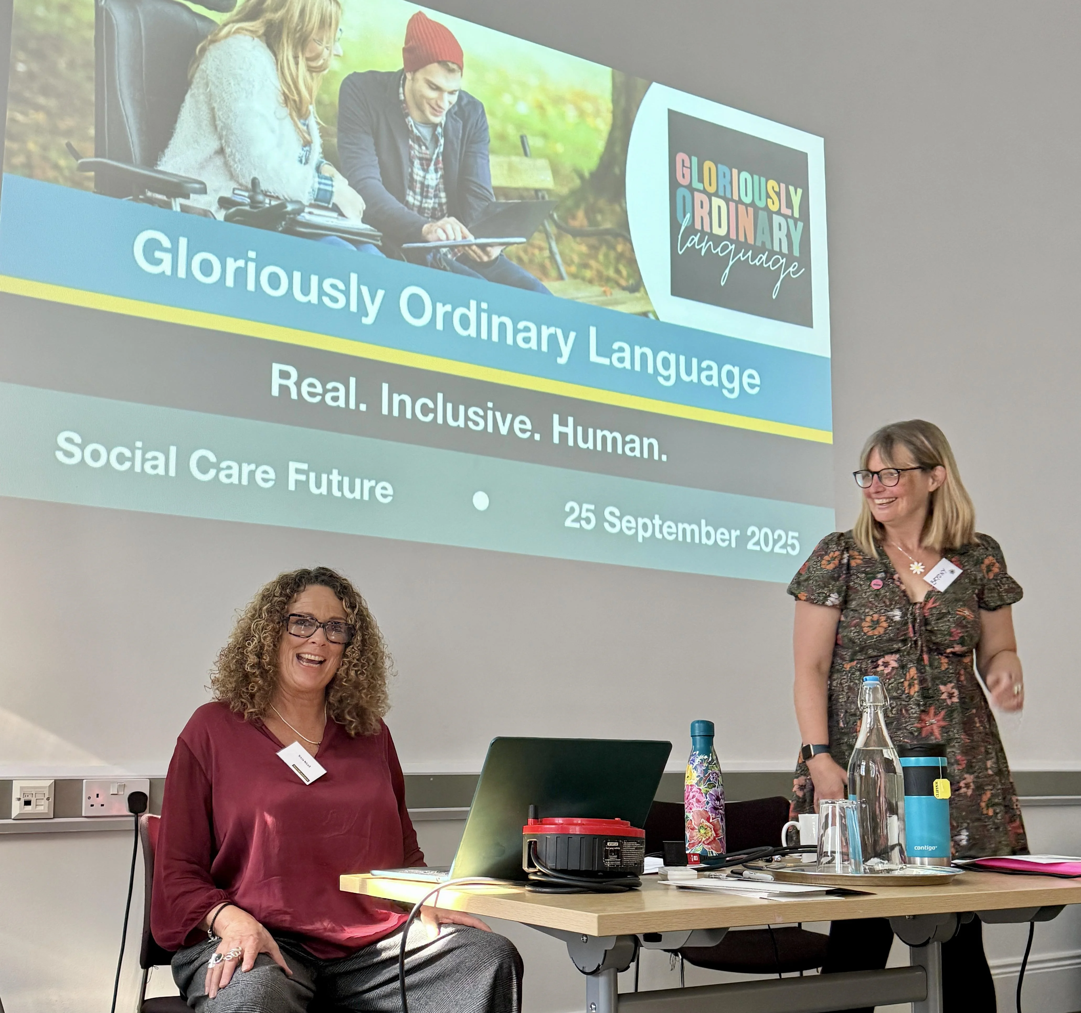 Two people at the front of a room presenting, standing and sitting behind a table with a laptop, water bottles, and papers. Behind them, a projected slide reads “Gloriously Ordinary Language: Real. Inclusive. Human.”