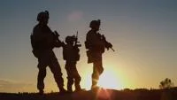 Three soldiers in battlefield gear silhouetted against the sun on the horizon.