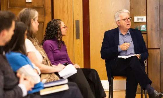 4 speakers are out of focus to the left of the image, a man in a suit on the right addresses a crowd who are off camera