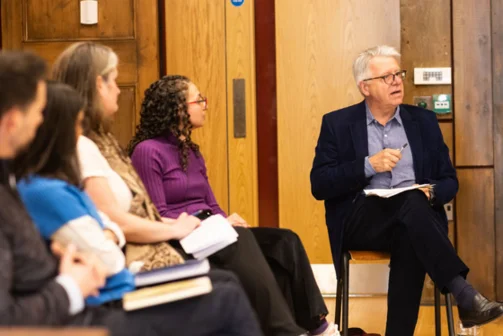 4 speakers are out of focus to the left of the image, a man in a suit on the right addresses a crowd who are off camera