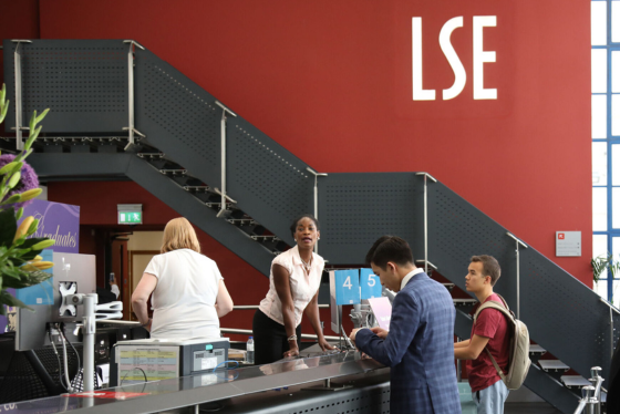 Students interacting with staff at LSE reception desk beneath distinctive red wall and staircase