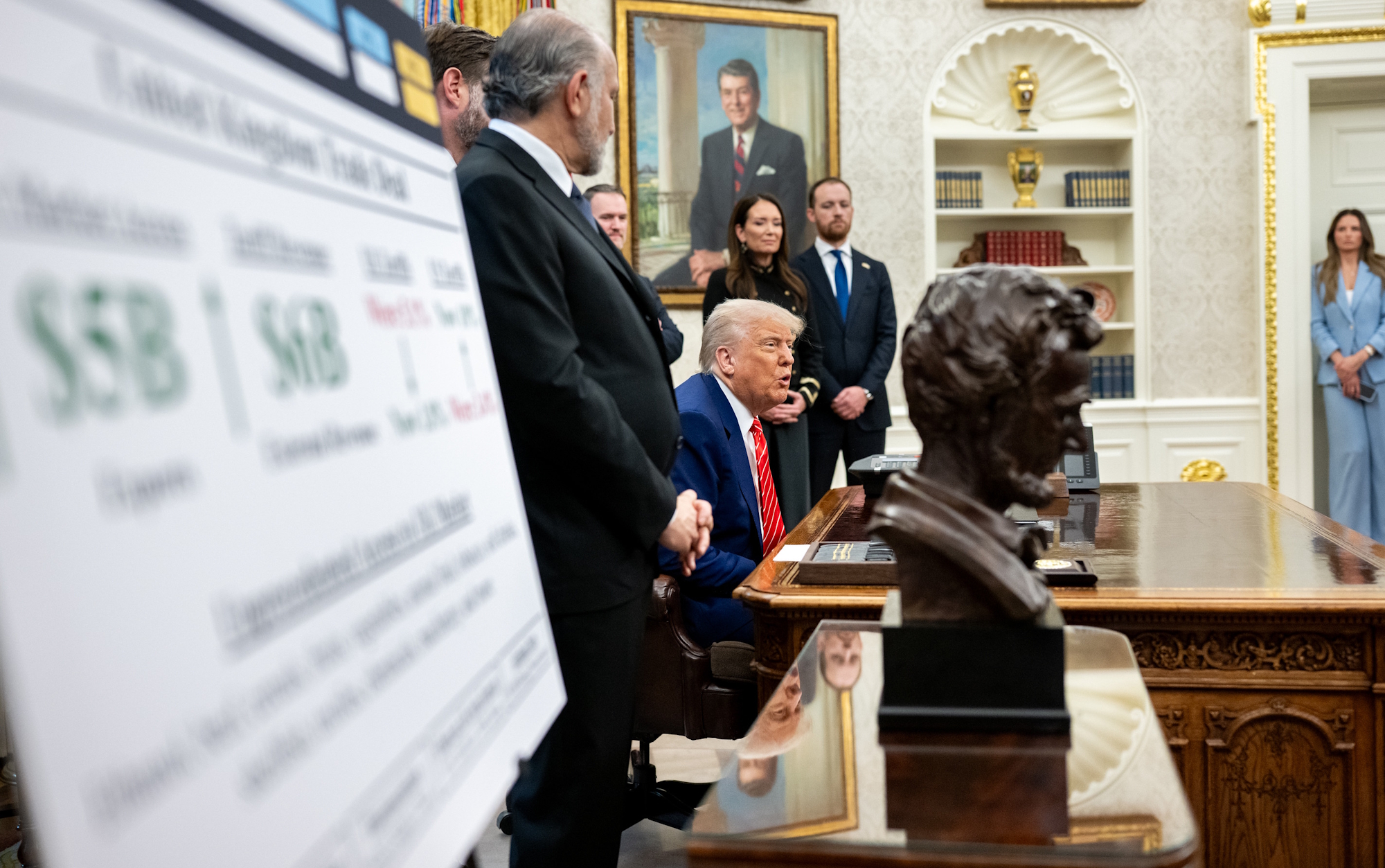 President Donald Trump seated at a large wooden desk, speaking, with aides standing behind him in a formal office setting.