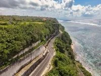 Elevated view of a curving road along a steep green cliff, turquoise ocean, with waves breaking along the shoreline