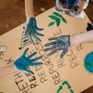 Paint-covered hands coming together over a cardboard sign with drawn symbols highlighting sustainability and recycling
