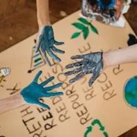 Paint-covered hands coming together over a cardboard sign with drawn symbols highlighting sustainability and recycling