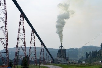 Industrial facility emitting a thick plume of smoke into the air near a large conveyor structure in a rural landscape