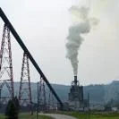 Industrial facility emitting a thick plume of smoke into the air near a large conveyor structure in a rural landscape