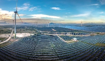 Expansive renewable energy site with rows of solar panels across rolling terrain, alongside multiple tall wind turbines