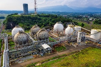 A large industrial energy facility with storage tanks and pipelines surrounded by green hilly landscape