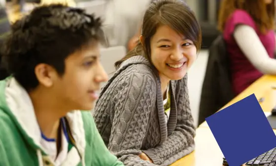 A student looks over at another student, smiling. Both are sat at a desk.