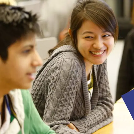 A student looks over at another student, smiling. Both are sat at a desk.