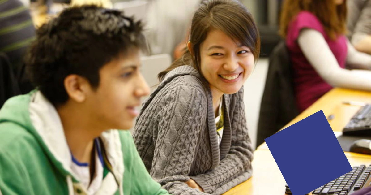 A student looks over at another student, smiling. Both are sat at a desk.