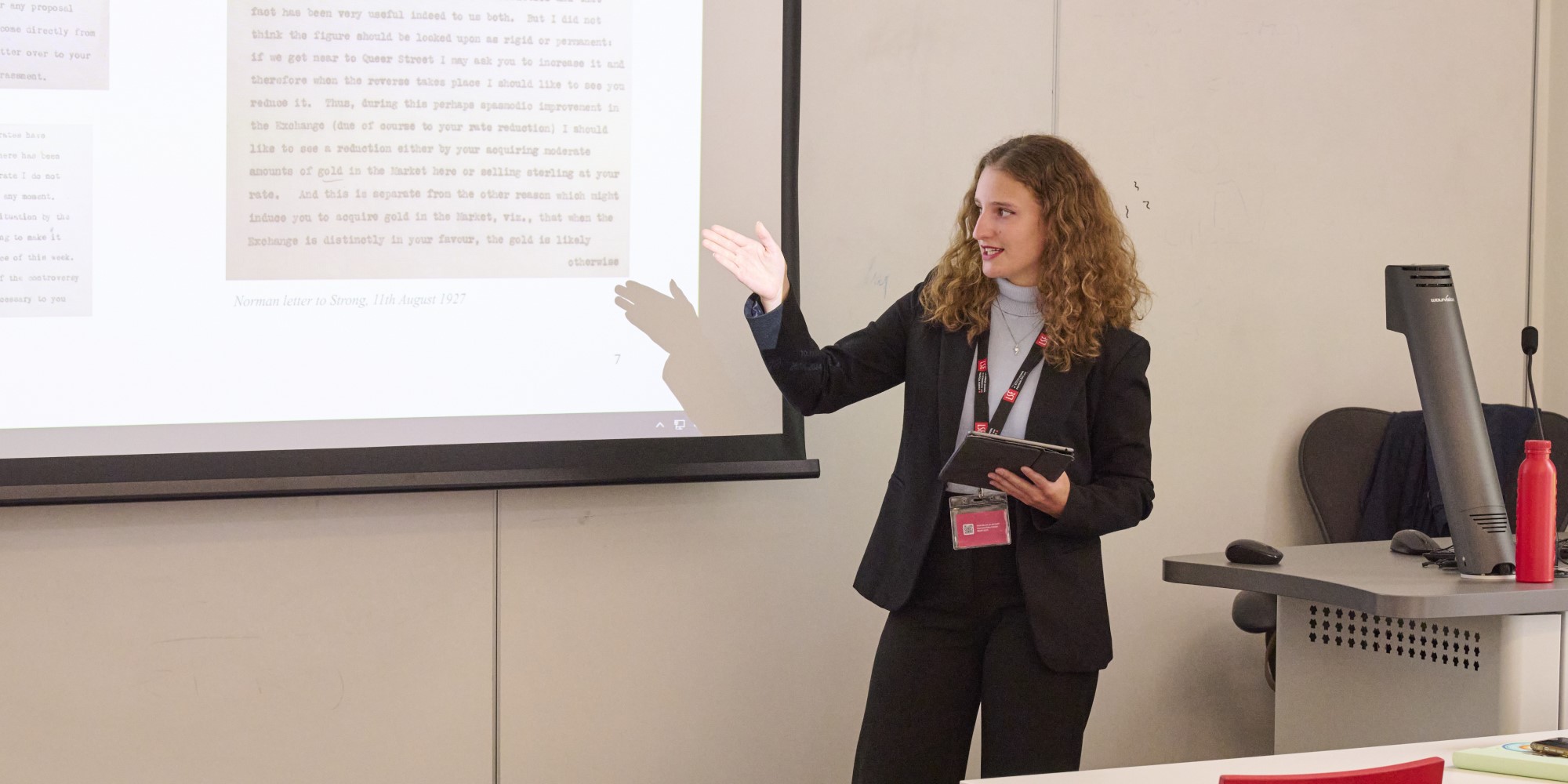 A speaker gesturing to a presentation at the front of a classroom