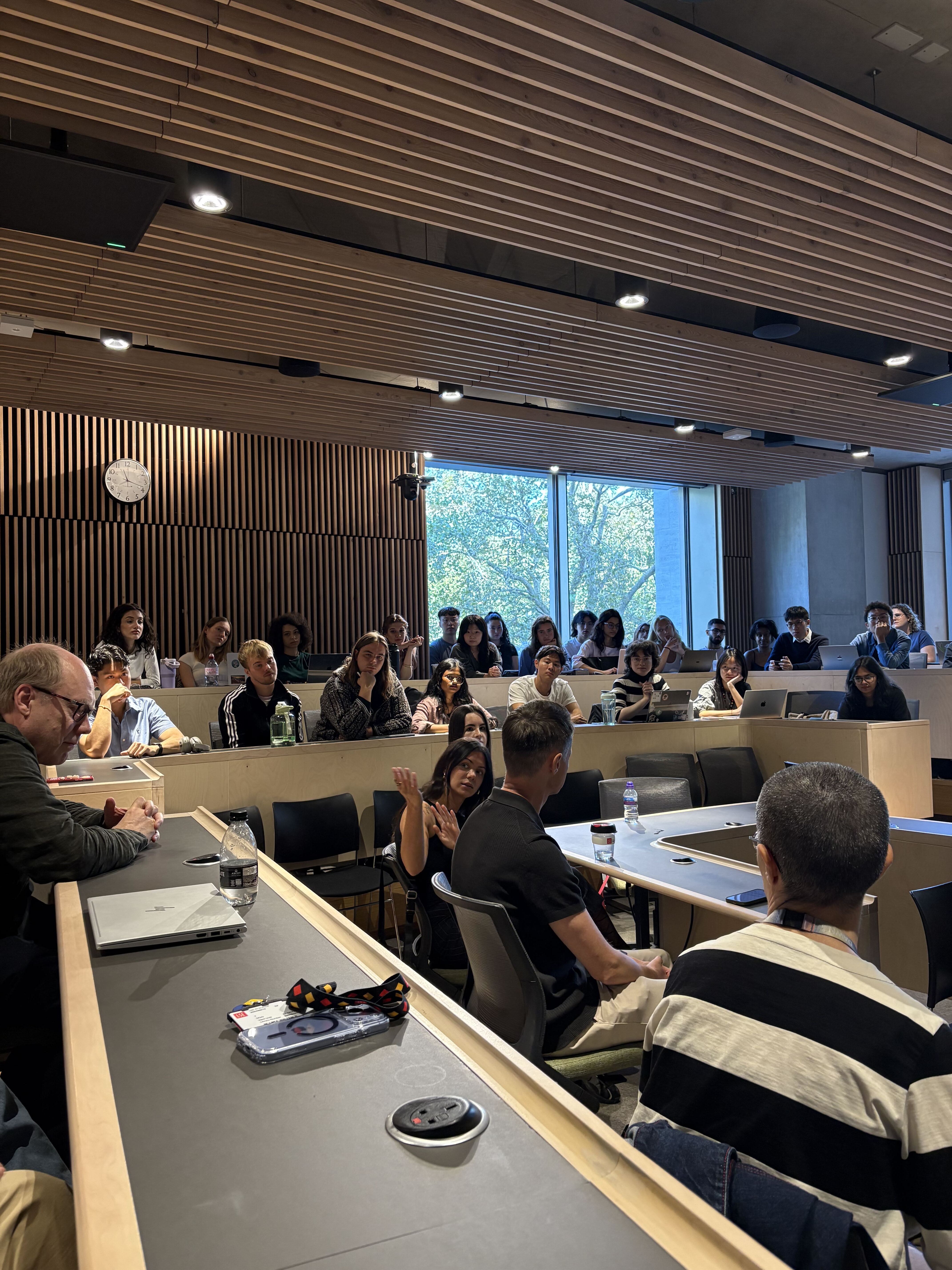 Students sitting at desks in a large, modern classroom or lecture hall with windows and wooden ceiling panels.