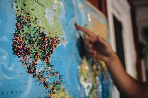 A hand pointing towards a wall map of North America with pins in the United States