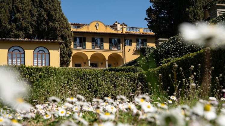 A historic yellow villa with arched colonnades and dark shutters, viewed across formal hedged gardens, white daisy flowers and tall cypress trees framing the building.