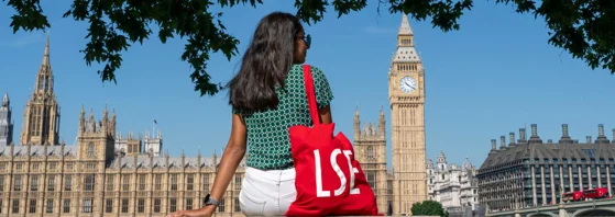 A woman sitting in front of Houses of Parliament with a red LSE tote bag