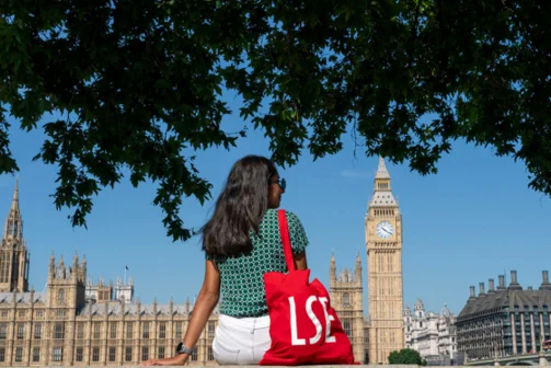 A woman sitting across from Houses of parliamnet with a red LSE tote bag