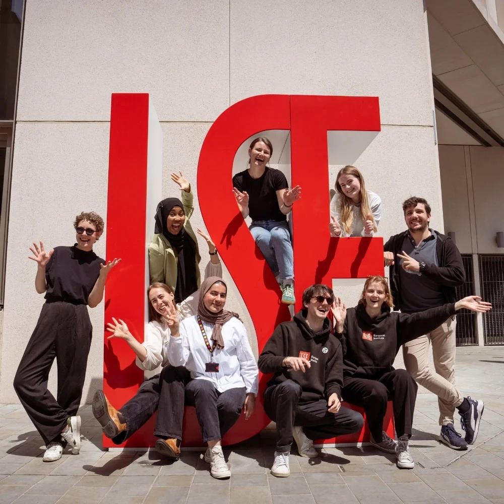 Students smiling and waving sat on large red letters spelling LSE