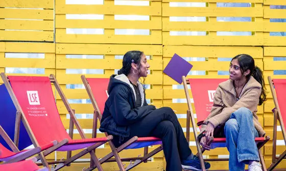 Two students sitting on red deck chairs chatting in front of a yellow backdrop