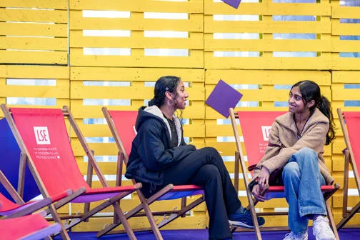 Two students sitting on red deck chairs chatting in front of a yellow backdrop