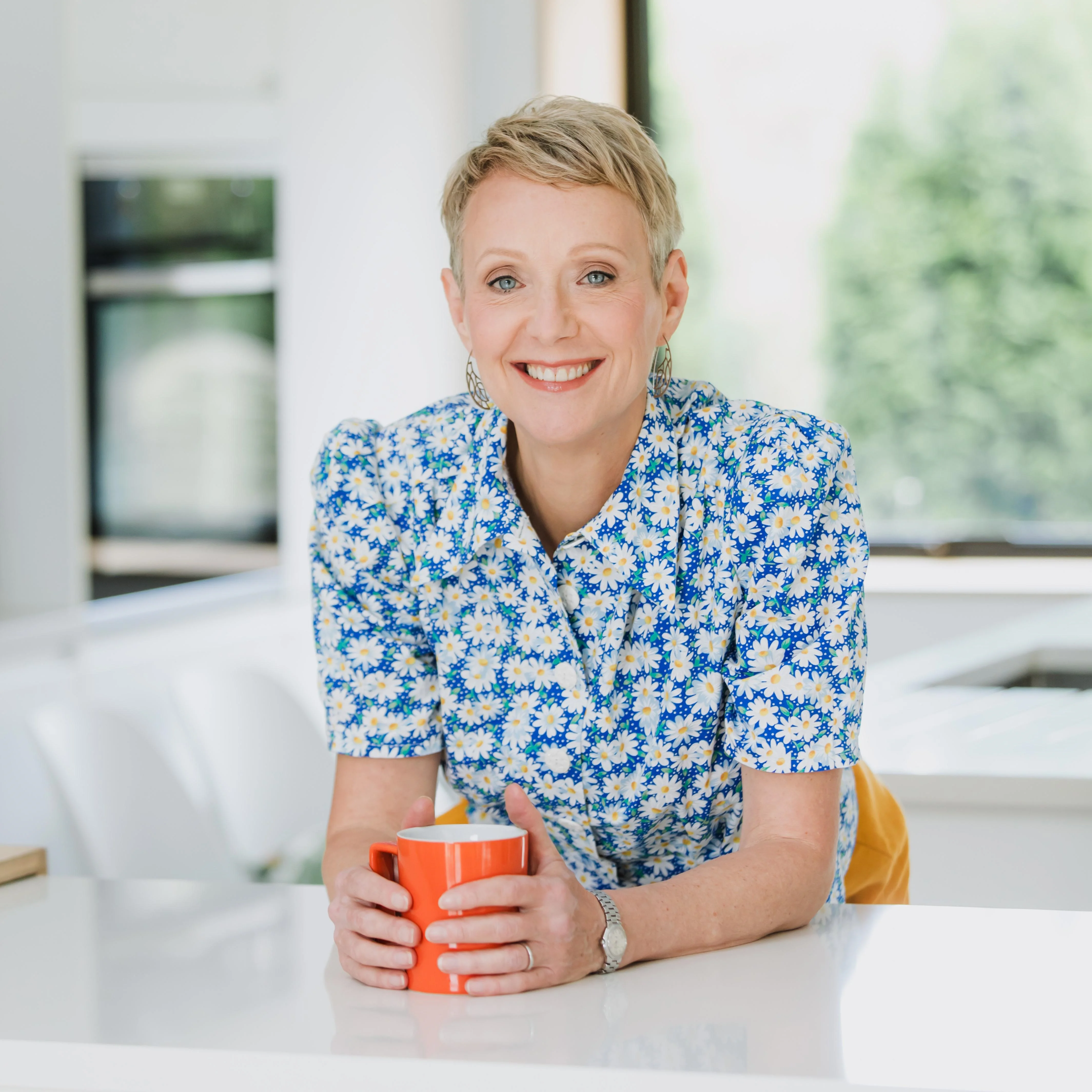 Wendy Lloyd is leaning on a kitchen workstop holding a orange mug in both hands. She is smiling and has a blue floral shirt on.