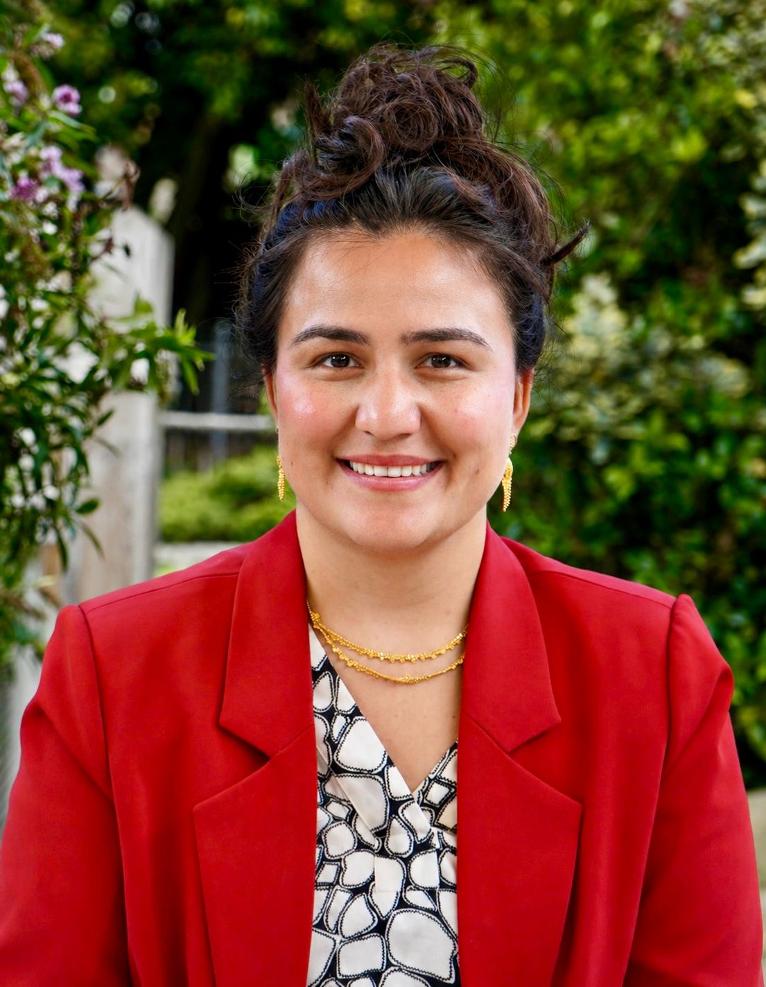 Rabia is smiling and facing the camera. She is wearing a red blazer with gold jewellery. 