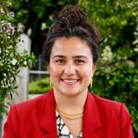 Rabia is smiling and facing the camera. She is wearing a red blazer with gold jewellery.