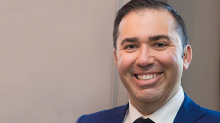 Brendan Gellman smiles at the camera. He is wearing a dark blue suit, white shirt and black tie.