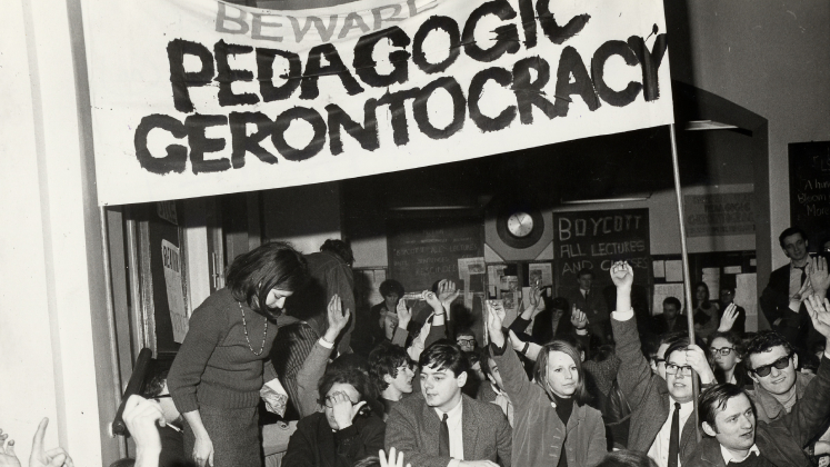 Black and white image of students gathering under a banner which says "beware the pedagogic gerontocracy"